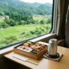 新幹線の窓際テーブルに並べられた美味しそうな駅弁と缶ビールの様子